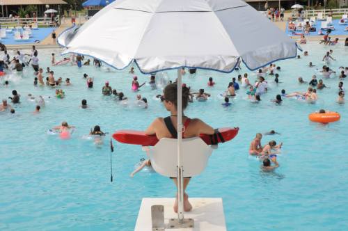 A lifeguard sits under an umbrella, overlooking a crowded swimming pool filled with people enjoying the water.