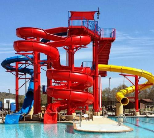 Colorful water slides in a pool area under a clear blue sky, featuring red and yellow slides and a tall observation deck.