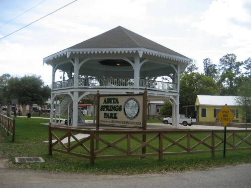 A gazebo in Arta Springs Park, surrounded by green grass and trees, with a sign at the entrance.