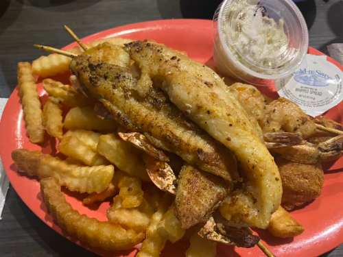 A plate of fried fish fillets, shrimp, and crinkle-cut fries, accompanied by a small container of coleslaw.