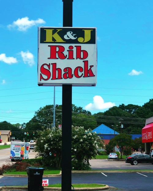 Sign for K&J Rib Shack with a blue sky and trees in the background, indicating the restaurant is open.