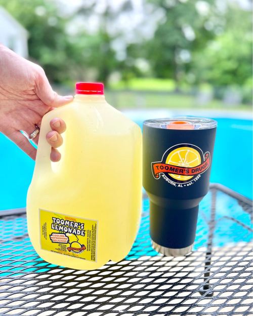 A hand holds a jug of lemonade next to a black cup with a logo, set against a blue pool background.