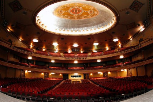 Interior of a grand theater featuring ornate ceilings and rows of red seats, illuminated by soft overhead lights.