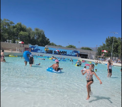 A busy water park scene with people swimming and floating in a pool under a clear blue sky.