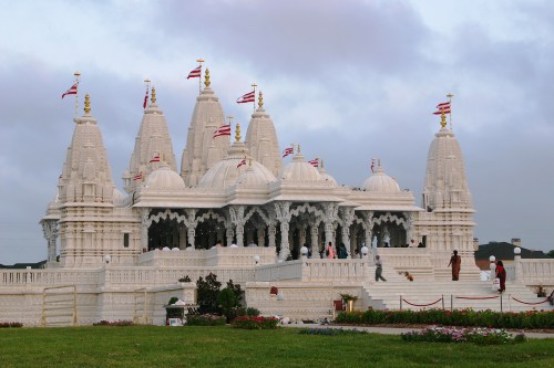 A grand white temple with intricate architecture, surrounded by greenery and flags, under a cloudy sky.