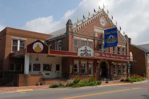 Historic theater building with a marquee, flags, and colorful flowers in front, under a partly cloudy sky.