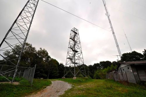 A gravel path leads to tall communication towers surrounded by trees under a cloudy sky.