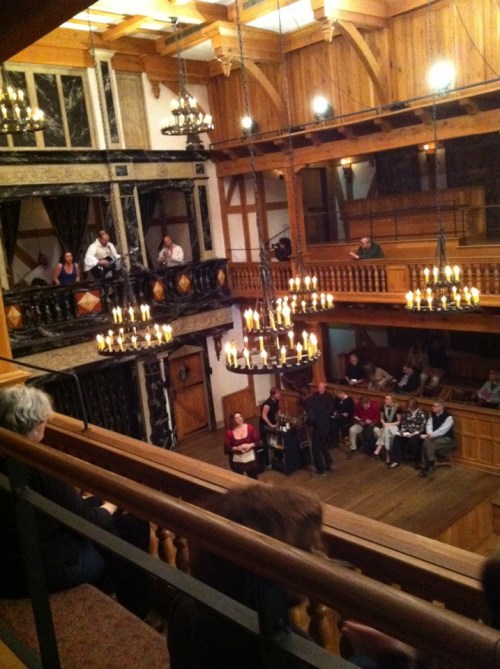 A wooden theater interior with chandeliers, featuring an audience seated in rows and on balconies, watching a performance.