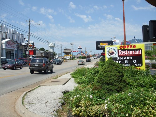 A busy street scene with cars, shops, and a restaurant sign advertising BBQ and steaks. Blue sky with scattered clouds.