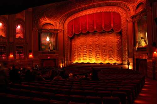 A dimly lit theater with ornate decor, featuring a red curtain and empty seats awaiting an upcoming performance.