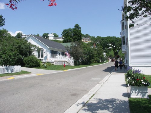 A quiet street lined with trees and houses, featuring an American flag and pedestrians walking on the sidewalk.
