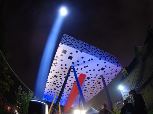A large, abstract structure with a patterned top is illuminated by a beam of light at night, surrounded by people.