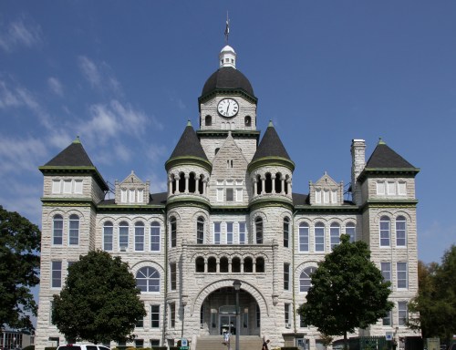 Historic stone building with a clock tower, featuring ornate architecture and green accents against a blue sky.