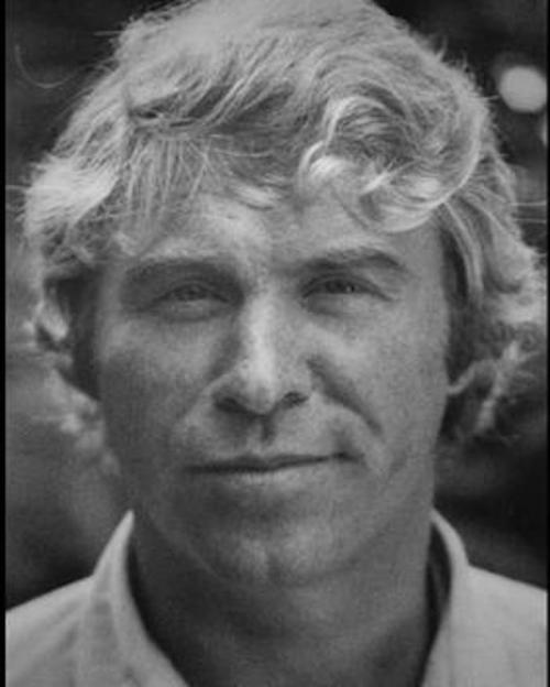 Black and white portrait of a man with wavy blonde hair, looking directly at the camera with a neutral expression.