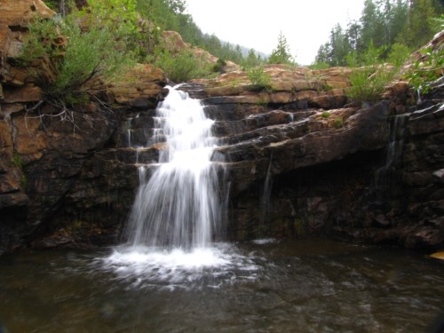 A serene waterfall cascading over rocky terrain into a calm pool, surrounded by lush greenery and forested hills.