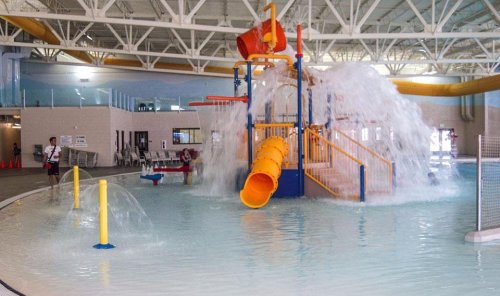 A colorful water play structure with slides and fountains in an indoor pool area. Water splashes around play equipment.