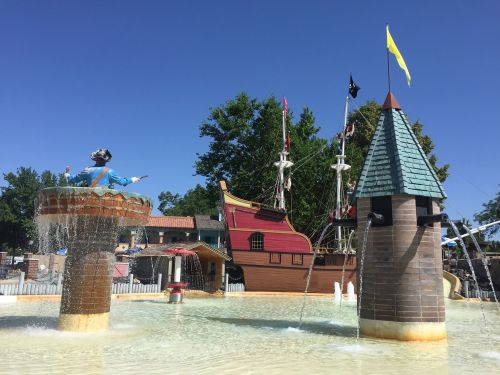 A playful water fountain with a pirate ship and castle in the background, under a clear blue sky.