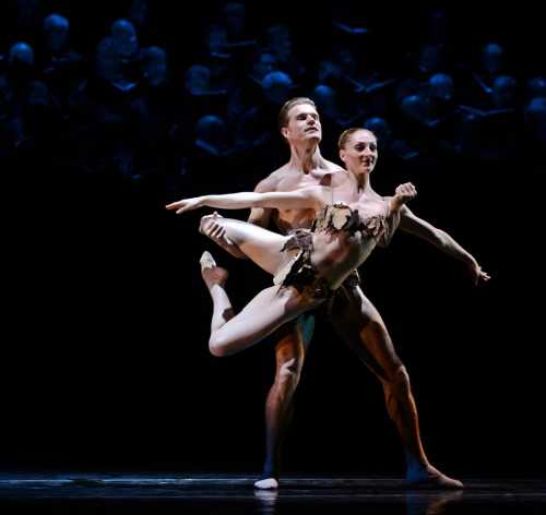 A male and female dancer perform a graceful ballet duet on stage, surrounded by a dark, shadowy audience.