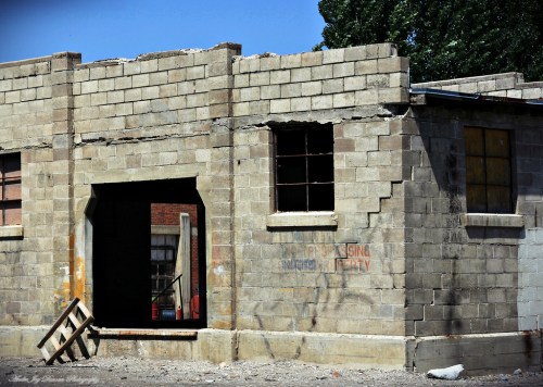 Abandoned building with a partially collapsed wall and an open doorway, surrounded by rubble and greenery.