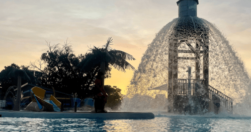 A water park scene at sunset, featuring a large fountain and a water slide with palm trees in the background.