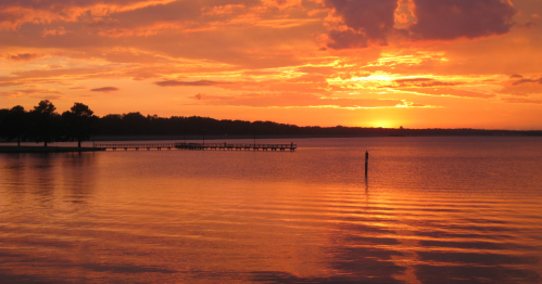 A serene lake at sunset, with vibrant orange and yellow hues reflecting on the water and a distant pier.