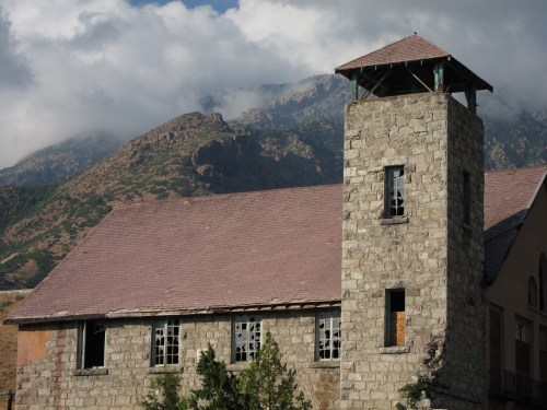 A stone building with a tower, set against a backdrop of mountains and cloudy skies. Windows are broken and overgrown.