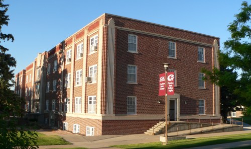 A brick building with multiple windows and a ramp, featuring a banner on a lamp post. Clear blue sky in the background.