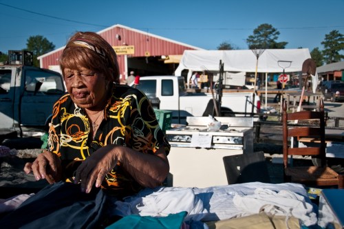 An elderly woman sorts through clothes at a bustling outdoor market, with various items and vendors in the background.