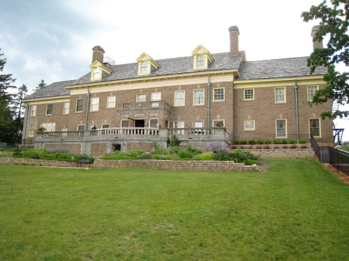 A large, historic brick mansion with yellow accents, surrounded by green grass and landscaped gardens.