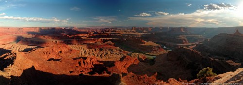 Panoramic view of a vast canyon landscape with red rock formations and a winding river under a blue sky.