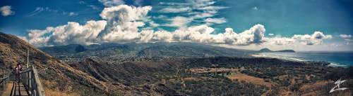 A panoramic view of a lush landscape with mountains, clouds, and a coastline under a bright blue sky.