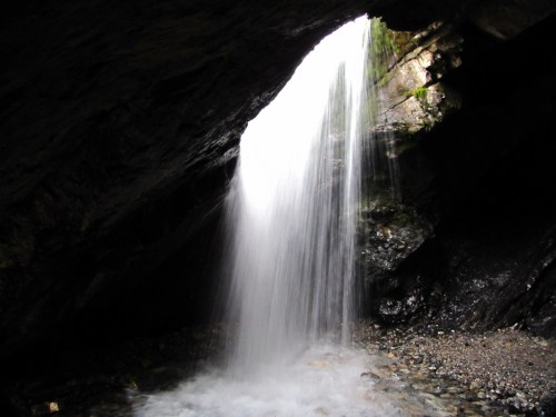 A waterfall cascading through a rocky cave, with sunlight illuminating the water and surrounding stones.