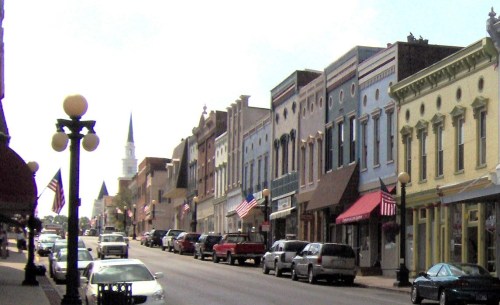 A charming small-town street lined with colorful storefronts, parked cars, and a church steeple in the background.