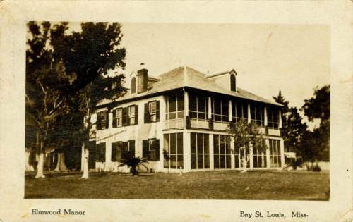 Historic Elmwood Manor in Bay St. Louis, Mississippi, featuring a two-story structure with a porch and surrounding trees.