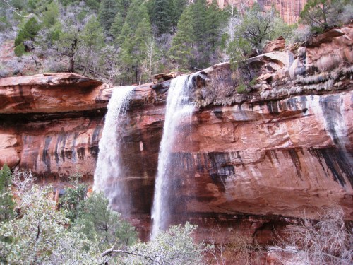 A waterfall cascading over red rock cliffs, surrounded by green trees and rugged terrain.