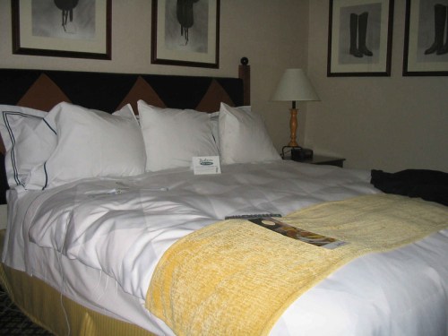 A neatly made bed with white linens and a yellow throw, in a cozy hotel room with framed artwork on the walls.