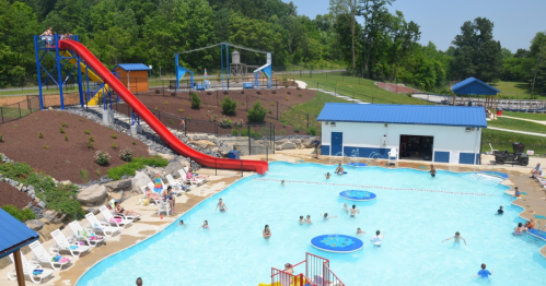 A sunny outdoor pool area with a water slide, people swimming, and lounge chairs around the pool. Green trees in the background.