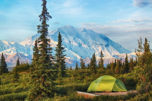 A green tent set against a backdrop of snow-capped mountains and lush trees under a clear blue sky.