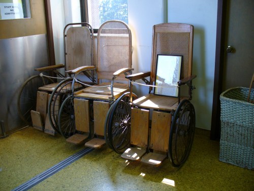 Two vintage wooden wheelchairs with caned seats are positioned near a window, casting shadows on the floor.