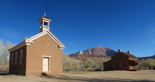 Historic adobe building and a wooden house against a clear blue sky, with mountains in the background.