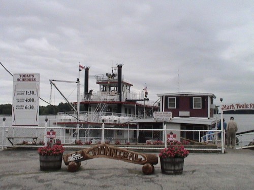 A riverboat docked at a pier, with a schedule sign and flower planters in the foreground under a cloudy sky.