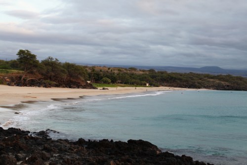 A serene beach at dusk, with gentle waves lapping at the shore and rocky formations in the foreground.