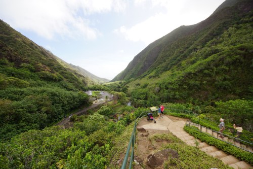 A scenic valley with lush greenery, a winding path, and people walking along the trail near a river.