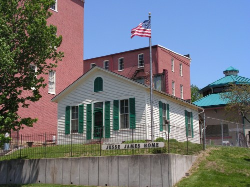 Historic Jesse James Home with green shutters, American flag, and surrounding greenery on a clear blue sky day.