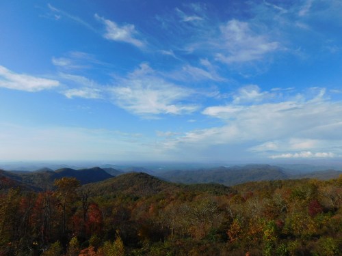 A panoramic view of rolling mountains covered in autumn foliage under a blue sky with scattered clouds.