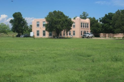 A two-story, light-colored building surrounded by green grass and trees under a blue sky.