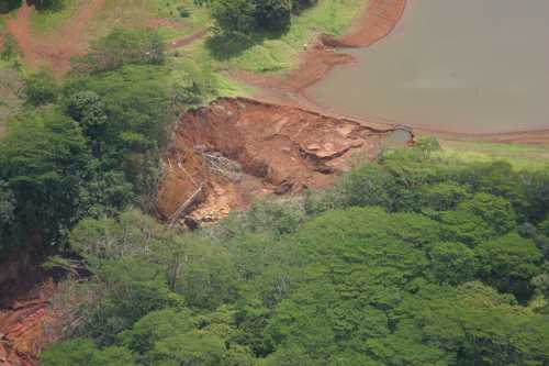 Aerial view of a landslide area with exposed soil and trees, near a body of water surrounded by lush greenery.