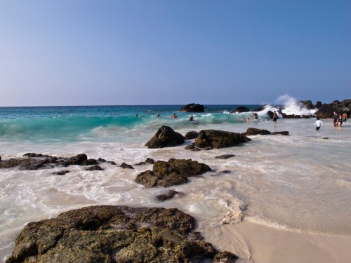 A scenic beach with turquoise waves, rocky formations, and people enjoying the water under a clear blue sky.