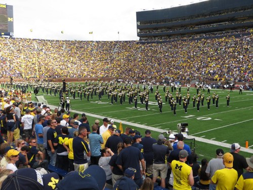 A large crowd watches a marching band perform on a football field at a stadium filled with fans in blue and yellow.
