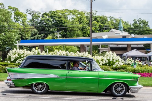 A classic green and black vintage car drives past a gas station, surrounded by lush greenery and flowers.